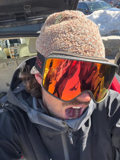 A close-up outdoor portrait captures a male individual in winter ski attire, located in a parking area in La Salle-les-Alpes, France. The subject wears a textured, light brown and off-white knit beanie, affixed with a gold-colored ski-themed pin on the right side. His upper face is obscured by large, orange-red mirrored ski goggles branded "ZEISS" with a "SPORT DESIGN" strap. His mouth is wide open, revealing his upper and lower teeth and tongue, suggesting an expressive vocalization or a gasp. He is dressed in a dark grey or black winter jacket, partially zipped, with the "Performance" brand visible on the lower right. Behind the subject, the open trunk of a light-colored vehicle, revealing its interior, is visible. The background includes piles of snow and other parked vehicles under bright daylight conditions.