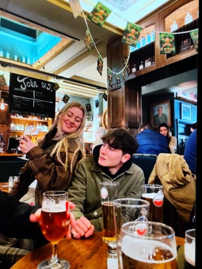 This image depicts an interior scene within a pub or bar in Braine-l'Alleud, Belgium, during a St. Patrick's Day celebration, indicated by festive green banners featuring leprechauns and "St. Patrick's Day" text strung across the ceiling.
In the foreground, a young adult female with long blonde hair, wearing a brown jacket, stands slightly off-center left, smiling and looking towards her left. She holds a stemmed glass containing an amber beverage in her right hand. To her right, a young adult male with dark, curly hair, wearing an olive-green jacket over a dark hoodie, is seated at a wooden table, observing the female. His hands are clasped on the table.
On the wooden table in front of the male and partially obscuring the view of the female, several glasses of beer are present. One stemmed glass contains amber liquid and shows a partial "EL AL..." logo. Two other tall, clear glasses, one mostly empty and one partially filled with amber liquid, bear "Amstel" branding. Another glass, partially visible in the extreme foreground right, also contains amber liquid. A salt or pepper shaker is also on the table.
In the mid-ground and background, the establishment features traditional pub decor with dark wood paneling and shelves stocked with bottles and glasses behind a bar area. A blackboard sign reads "Join us." and lists "COCKTAILS" and other menu items. A framed portrait of an older man hangs on a wall in the background. Several blurred individuals are visible in the background, including one person in a blue puffer jacket, indicating other patrons are present. The lighting is warm and ambient, characteristic of a pub environment.