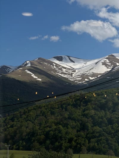 The image displays a mountain landscape with a snow-covered peak on the left and a densely forested area on the right. A bright blue sky with scattered clouds fills the upper left corner. Power lines are visible, cutting across the scene from the upper right to the lower center. The image is rotated 90 degrees clockwise, making the sky appear on the left and the ground on the top. There are no people visible in the image. Based on the location provided (Lermontovo, Armenia), the mountain is likely part of the Pambak Mountain Range.