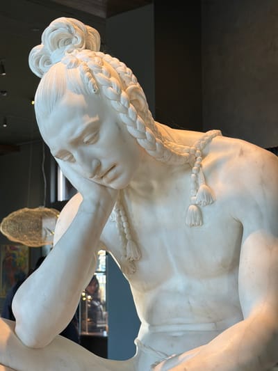 A close-up captures a white marble sculpture, likely depicting a Greek or Roman figure. The head is inclined with eyes closed. Braided hair adorns the head and is wrapped around the body with tassel accents. The sculpture appears to be in a museum or gallery setting, with a dark wall behind it. Reflections hint at other artworks in the vicinity. The lighting emphasizes the textures and contours of the sculpture.