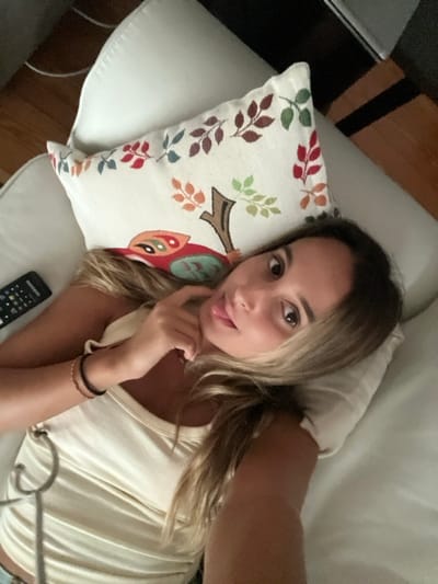 A young adult female is depicted lying on her back on a light-colored upholstered surface, likely a couch or bed, within an indoor setting in Oliveira de Frades, Portugal. She has long, light brown hair with blonde highlights, fanned out around her head. Her brown eyes are open and directed towards the camera. Her lips are slightly parted, revealing the tip of her tongue. Her right hand (viewer's left), adorned with a dark and a light-colored bracelet on the wrist, has its fingers positioned near her chin. She is wearing a light-colored, ribbed tank top with a silver-colored, chain-like embellishment visible on the left side. Her head rests on a small, light-colored pillow. To her left (viewer's right) on the upholstered surface, a larger, off-white decorative pillow is present, featuring an embroidered design of autumnal leaves and a stylized red owl. A black television remote control is situated on the surface to the left of her head. The background consists of partially visible dark and light brown wooden surfaces.