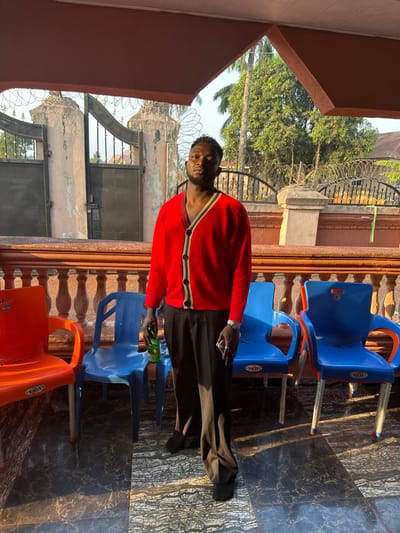 An individual in a red cardigan and black trousers stands outdoors beside stacked blue plastic chairs and an orange chair. They hold a green bottle. Behind them, a building with a fence topped with barbed wire and trees is visible. The scene is captured on a textured tile surface, with architectural elements of the building framing the view.