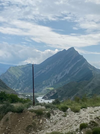 The image depicts a mountainous landscape with a river winding through a valley under a cloudy sky. In the foreground, a dirt path with scattered rocks and sparse vegetation leads towards the middle ground. A single, tall, dark pole with wires stretches across the scene, partially obscuring the view of the mountains and river. The mountains themselves are rugged and covered in green vegetation, with some rocky outcrops visible. The river appears to be flowing through a gorge, with lighter-colored water. The overall impression is of a natural, somewhat remote, and undeveloped area. The metadata indicates the location as Chalda, Russia.