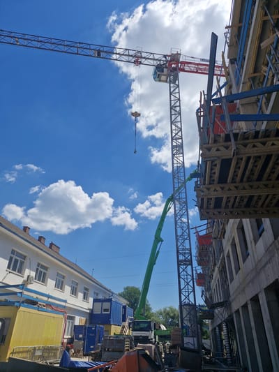 This media file captures a construction site with two prominent tower cranes against a bright blue sky dotted with white clouds. The image is oriented vertically, with the sky filling most of the frame.

The main subjects are the industrial machinery. The larger, red-and-grey tower crane is in the foreground, its boom extending diagonally upwards into the sky. The cab of the crane is visible. A second, darker metal crane structure is positioned behind and to the right of the first, its latticework reaching across the frame.

In the background, a multi-story building with white walls and several windows is visible. To the right of this building, several blue shipping containers and a yellow structure are present, suggesting temporary structures or storage units on the construction site. Some trees are also visible in the distance.

The scene appears to be set during daytime, given the bright sunlight and the clear blue sky, which suggests fair weather. The presence of construction equipment indicates an ongoing construction project. There is no visible text in the image. The context provided indicates the location is Brno, Czechia.