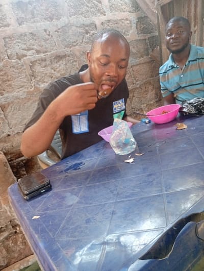Two men sit at a blue plastic table. One man eats from a spoon, eyes closed, wearing a black shirt. A pink bowl and a water bottle are nearby. The other man, wearing a blue and white striped shirt, watches. A lighter and remnants of something are also on the table. A black, rectangular object sits on the table's corner. The background features a brick wall and a glimpse of corrugated metal.