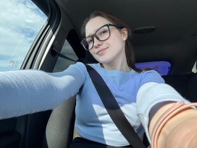 A woman wearing glasses and a light blue ribbed shirt is sitting in a car, wearing a seatbelt. The car's interior is visible, including a sunroof and the dashboard. The location is not identifiable as Zanevka, Russia from the image.