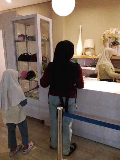The image shows three people in a room in a building in Kecamatan Margorejo, Indonesia. The room is lit by ceiling lights and a lamp. The room has white walls and a white tiled floor. There is a glass display case containing various items. There is a white counter with a blue rope barrier. One of the people, wearing a grey hooded garment, is standing by the counter. The other two people are facing the counter. The person standing next to the counter is wearing a black hijab. They are holding a black backpack with a red strap. The person behind them is wearing a white hijab and a long-sleeved white shirt. The person behind the counter is wearing a white hijab and a long-sleeved white shirt and has dark hair. They are standing on the left side of the counter and appear to be interacting with the people in front of them.