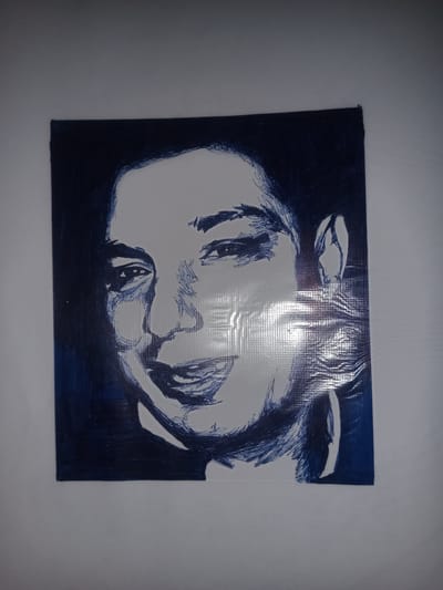 The image displays a sketch of a person's face drawn in dark blue ink on a white surface. The drawing is centered on a sheet of material with a slightly textured, possibly plastic, surface. The sketch depicts a close-up of the face, with the left side of the face shaded more heavily. The eyes are detailed, and the mouth is open as if speaking or smiling. There is a noticeable shine and a grid-like pattern visible on the surface, suggesting it might be a laminated poster or banner. The drawing is bordered by a dark blue background. The image does not contain any information about the city of Gulbag, Uzbekistan.