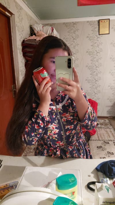 A young woman with long, dark hair holds a red soda can in her right hand while taking a selfie with her smartphone in her left hand. She is wearing a dark blue dress with a floral pattern of red and white flowers. Her left hand is adorned with a ring. The background appears to be a domestic setting with floral wallpaper and a mirror reflecting the scene. The woman's face is partially obscured by the can and the phone. On a table in the foreground, several items are visible, including what appears to be a container of petroleum jelly and other unidentifiable objects. The scene is lit by artificial light.