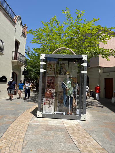 The image depicts a street scene in La Roca del Vallès, Spain, likely at an outdoor shopping village. A large, transparent display case, resembling a trunk, stands prominently in the foreground, showcasing a mannequin dressed in an outfit of blue jeans and a green top, along with various retail items and imagery. In the background, people are walking along a paved walkway. On the left, two men and a woman are walking towards the display. To the right, another woman in a red top and white pants is walking away from the camera. The buildings flanking the street have traditional architecture with balconies and arched doorways. Lush green trees with bright leaves overhead provide shade. The sky is clear and blue.
