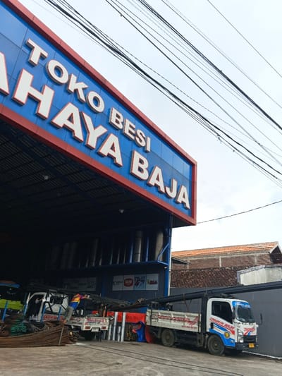 A building with a blue and red sign reading "TOKO BESI JAYA BAJA" dominates the foreground. Underneath the sign, trucks are parked, seemingly being loaded with metal materials. Electrical wires crisscross the upper portion of the frame against a pale sky. The rear of the building and adjacent structures are visible in the background.
