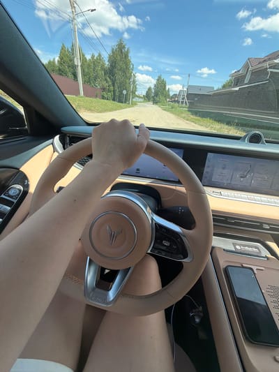 A first-person perspective captures a person driving a car on a sunny day, with the exterior view visible through the windshield. The scene appears to be a rural road, likely in Malinovka, Russia, judging by the presence of wooden fences, modest houses with pitched roofs, and lush green trees lining the roadside. The road itself is unpaved and dusty. Inside the car, the steering wheel, dashboard, and a large touchscreen display are prominent. The driver's right arm is visible, resting on the steering wheel. A smartphone is placed on the center console, indicating it's within the car's cabin. The overall impression is one of a casual drive through a suburban or rural setting.