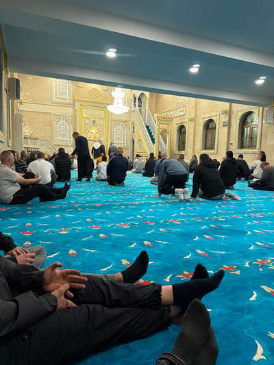 The image displays the interior of a mosque located in Breznitsa, Bulgaria. Approximately 20 to 30 adult males are present, predominantly seated or kneeling on a vibrant blue carpet adorned with repeating white, orange, and yellow patterns. Several individuals are positioned in prayer postures, while others sit casually, some with legs extended. Attire consists of various casual clothing including trousers, shirts, hoodies, and jackets; most are wearing socks or are barefoot.
The architectural features include light-colored walls extensively decorated with intricate gold-colored moldings and panels. Centered at the front is an ornate mihrab, a prayer niche, accented in gold. To the right of the mihrab, a white and gold minbar, a tiered staircase pulpit, is visible. The right wall contains several arched windows with dark frames and decorative grilles. The ceiling is light blue, fitted with recessed spotlights, and features two multi-tiered crystal chandeliers providing illumination. In the foreground, the lower bodies and legs of two individuals are visible, seated on the carpet, one with hands clasped, both wearing dark socks.