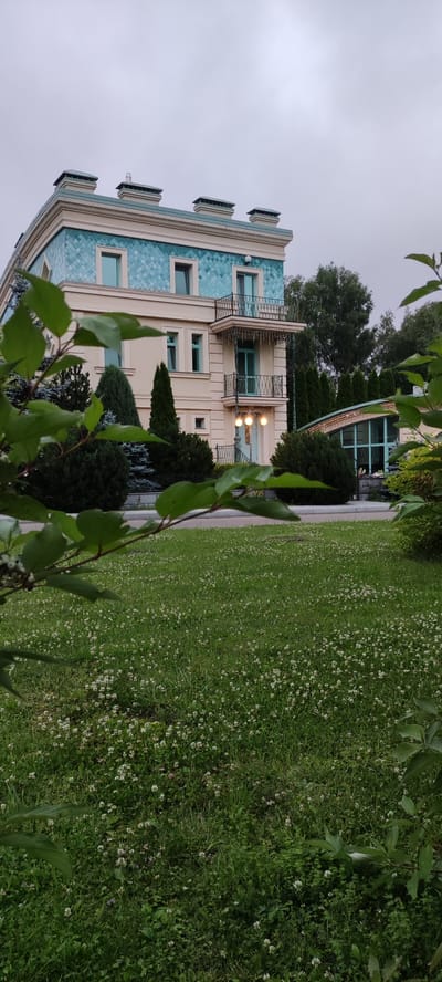 This image shows a multi-story building with a light cream facade and a distinctive turquoise tiled roof and upper wall section. The building has multiple windows and balconies with black wrought-iron railings. In front of the building is a well-maintained lawn covered with small white flowers, possibly clover. Several evergreen trees and bushes are planted around the building. To the right of the main building, a smaller structure with a curved glass roof is partially visible. The sky is overcast, suggesting a cloudy day. The image appears to be taken in the grounds of the Ангелово Клубная Резиденция in Russia. No people are visible in the image.