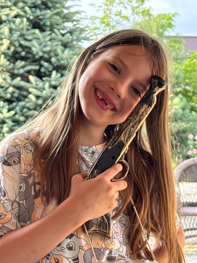 In this close-up, slightly tilted, outdoor photograph, a young girl with long brown hair is holding a toy gun. She is smiling broadly at the camera, revealing a gap where a tooth is missing. Her hair is brushed back from her face, with some strands falling forward. She is wearing a light-colored shirt with a repeating pattern of cartoon cats. The toy gun has a brown and black camouflage design.

The background is blurred, but appears to be a backyard setting with green foliage, possibly a tree or bushes, and hints of a building. The lighting suggests it is daytime, likely a sunny day. The girl's expression is joyful and playful. There is no visible text in the image. The location context is Zassovskaya, Russia.