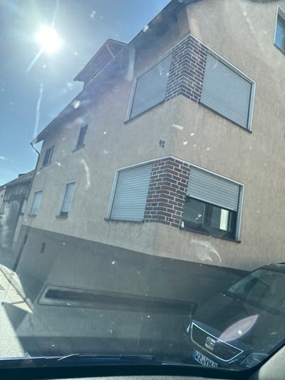The media file displays a reflection in a car windshield, showing a modern building with several windows and roller shutters. The building appears to be a residential or commercial property with a textured, light-colored facade. Some windows are open, revealing the interior, while others are covered by grey, ribbed roller shutters. Decorative brickwork is visible beneath some of the shutters.
The reflection also shows the sky, which is bright blue with some sunlight, suggesting it is daytime. The sun's glare is prominent, creating lens flare effects. The scene is likely a street view, given the perspective from inside a vehicle. The license plate of a vehicle is visible in the reflection, showing the text "WZ-YM9". A number "92" is faintly visible on the building's facade. The overall impression is a sunny day in an urban or suburban environment. The angle of the shot and the reflections suggest the photo was taken while driving or parked, looking at a building across the street. There are no people clearly visible in the media.