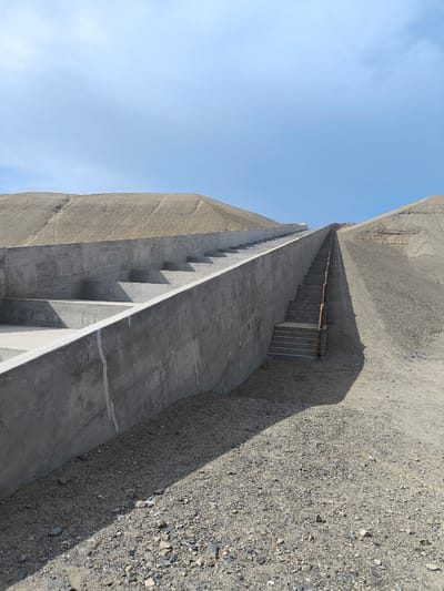 The image shows a long, steep staircase ascending a sandy hill in Huanchaco, Peru. The staircase is constructed of concrete and wood. The bottom of the stairs has a concrete structure with several levels. The surrounding landscape is arid and dusty with low hills on either side. The sky is clear and blue. No people are visible in the image.