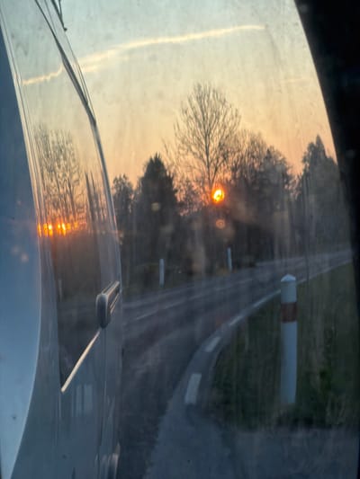 The image, captured from within a vehicle in Le Saulcy, France, presents a rural road scene during either sunrise or sunset. No individuals are present. The left foreground is occupied by the light-colored side body panel of the vehicle, featuring a dark-colored door handle and reflecting multiple instances of the bright orange sun and surrounding bare trees. The view through the vehicle's streaky window reveals a paved road, marked with a white dashed line, curving sharply to the right. A grassy embankment lines the roadside, where several white bollards with orange bands are positioned. In the background, a dense line of mostly bare deciduous trees and some evergreen foliage is silhouetted against a sky displaying gradients of orange and yellow near the horizon, transitioning to light blue overhead. The sun, an intense orange-yellow disc, is low on the horizon, partially obscured by tree branches, and exhibits lens flare effects. A faint contrail is visible in the upper sky.