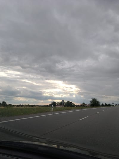 This image shows a view from a vehicle traveling on a paved road with a white line marking its left edge. The road curves gently to the right. On the left side of the road is a grassy field with some wildflowers and scattered trees. In the distance, a small town or village is visible, with several buildings and trees. The sky is overcast with heavy gray clouds, but there are breaks in the clouds where sunlight is shining through, creating a dramatic effect. The overall scene suggests a rural or suburban environment in the Czech Republic, possibly near Tábor, given the architecture and landscape. There are no people or animals visible in the image.