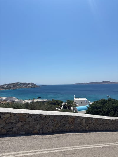 The scene depicts a panoramic view of the coastline in Agios Ioannis Diakoftis, Greece, under a clear, bright blue sky with no visible clouds. In the immediate foreground, a segment of an asphalt road with two parallel solid white lines runs horizontally. Above the road, a low, dry-stacked stone wall with a flat, light-colored coping extends across the frame. Beyond the wall, a verdant landscape features dense green foliage, including trees resembling olive trees and palm trees, partially obscuring white, cubic-shaped buildings with traditional Greek architectural elements. One prominent white building in the mid-ground features a small belfry or decorative structure on its roof and displays a Greek flag on a flagpole. This building also has a distinct, light blue curved or domed section. Further out, the Aegean Sea stretches across the mid-to-background, exhibiting shades from turquoise near the coast to deeper blue further offshore. Several boats are present on the water, including a dark vessel on the left and a white recreational boat on the right. In the distance, a landmass on the left is heavily populated with white buildings, while a larger, darker, undeveloped island or peninsula is visible on the far right horizon. No individuals are present.