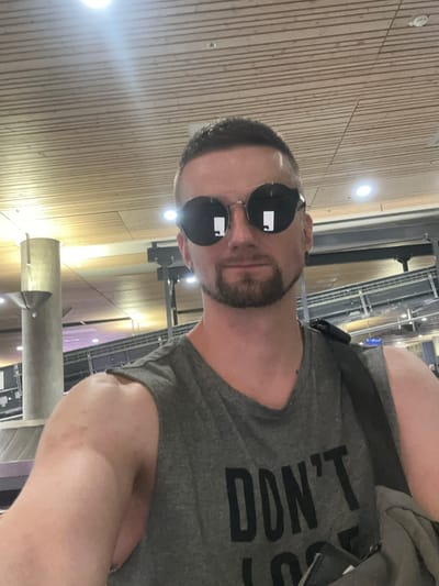 A man with short, dark hair and a goatee is taking a selfie indoors, likely at an airport in Gardermoen, Norway. He is wearing round, dark sunglasses with reflective lenses, a gray sleeveless t-shirt with the word "DON'T" printed in black letters, and a black backpack slung over his right shoulder. The background features a wooden-slatted ceiling with bright lights, a large concrete pillar with a metallic light fixture, and what appears to be baggage claim infrastructure with metal railings and a conveyor belt. The lighting suggests an indoor, public space with artificial illumination.