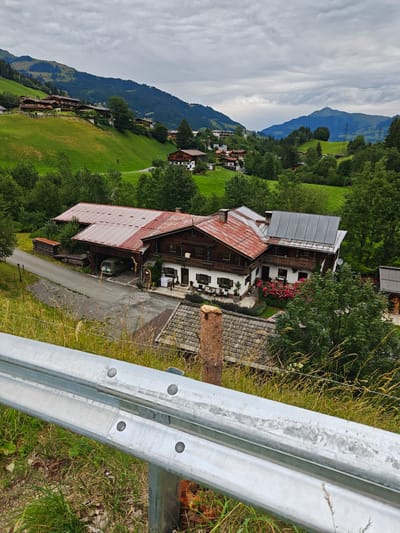 The image shows a rural landscape in Filzen, Austria. In the foreground, a silver guardrail runs horizontally across the bottom of the frame, with tall grasses and dry vegetation on either side. Behind the guardrail, a complex of buildings, likely a farm or guesthouse, is visible. The main building features a large, red, corrugated metal roof and a white facade with wooden balconies and shutters. A van is parked under a covered section of the building. In front of the building, a flowerbed with numerous pink roses is in bloom. The surrounding environment is characterized by rolling green hills covered in trees and some scattered houses. In the distance, mountains rise under a cloudy sky.