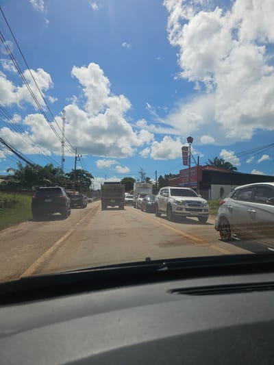 The image depicts a traffic scene on a rural road in Ban Thung Aok, Thailand, under a bright, cloudy blue sky. The road is paved with a yellow line marking the center and appears to have some dirt and gravel on the surface. Several vehicles are present, including a black SUV, a white SUV, a dump truck, and other cars, suggesting moderate traffic. Power lines and a tall telecommunications tower are visible along the side of the road, indicating the presence of infrastructure. In the background, there are buildings with signage, including what appears to be a business or shop. Trees and vegetation line the roadside, characteristic of a tropical environment. The photograph is taken from inside a vehicle, looking forward, as indicated by the dashboard at the bottom of the frame.