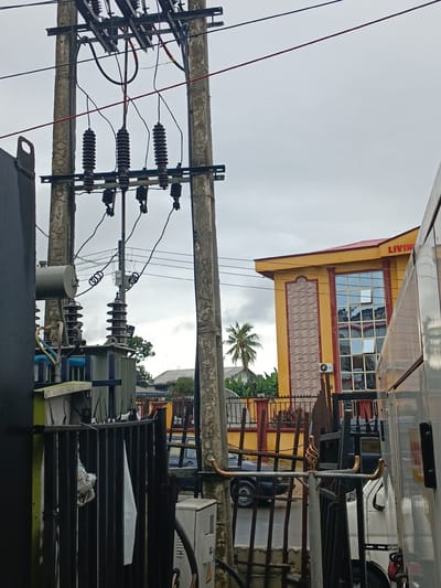 This image depicts an outdoor scene in Rumuokwuta, Nigeria, likely near an electrical substation. The foreground is dominated by utility poles with electrical insulators and wires, along with a large electrical transformer unit on the left. A black metal fence runs across the mid-ground, with some debris, including what appears to be white fabric, caught on it. Beyond the fence, a yellow building with red trim and large windows is visible. The building has the word "LIVING" in red letters on its facade. A palm tree and dense foliage are situated behind the fence and to the left of the yellow building. Several vehicles, including a black car and a white truck, are present in the background, suggesting a street or parking area. The sky is overcast, indicating a cloudy day. The overall impression is an urban environment with visible electrical infrastructure and commercial or residential buildings.