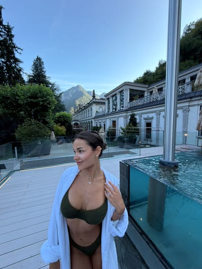 A woman in a dark green bikini and a white robe stands on a wooden deck next to a glass-enclosed pool. The woman is looking to her left, with her right hand resting on her hip. Behind her, a large white building with ornate architectural details is visible, nestled against a lush green hillside. Trees are scattered throughout the landscape, and mountains rise in the background under a clear blue sky with a crescent moon. The scene appears to be at a resort or spa in San Pellegrino Terme, Italy.