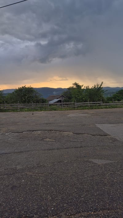 The image depicts an outdoor scene with a focus on a rural landscape and a damaged road in the foreground. In the distance, rolling hills are visible under a cloudy sky, with a band of warm-toned light, likely from a sunset or sunrise, breaking through the clouds near the horizon. A weathered wooden fence runs horizontally across the middle ground, separating a grassy area from a line of trees and a small, rustic building with a corrugated metal roof. The building appears to be a barn or shed. The foreground is dominated by a cracked and worn asphalt road, with visible potholes and uneven surfaces. A single black bird is perched on the road in the lower left quadrant of the image. The overall impression is one of a remote, possibly agricultural, setting experiencing inclement weather. The location is identified as Pletena, Bulgaria.