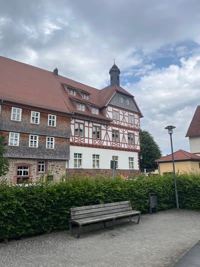 This image depicts a view of a historic building with traditional German half-timbered architecture, likely located in Neustadt, Germany. The building features a red tiled roof and a prominent dark-colored spire. White framed windows are visible across its facade, with some sections displaying the characteristic wooden beams of half-timbering. The building appears to be multi-storied, with different sections showing variations in architectural style and materials, including stone at the lower levels.

In the foreground, a paved walkway with a textured pattern runs alongside a dense, well-maintained green hedge. A weathered wooden bench with dark metal legs is positioned on the walkway, adjacent to the hedge, inviting rest. A modern street lamp with a cylindrical light fixture stands near the hedge and walkway. A metal trash bin is also visible near the bench and hedge.

The sky is partly cloudy, with a mix of bright white clouds and patches of blue sky, suggesting it's daytime. The image is taken from a slightly elevated perspective, looking upwards at the building and sky. There are no people visible in the frame, giving the scene a tranquil and unoccupied feel. The overall impression is one of a picturesque historical town setting on a pleasant day. No discernible text is present in the image.