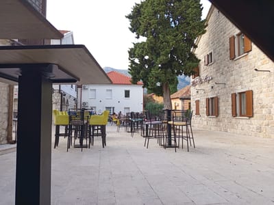This is an outdoor shot of a seating area at what appears to be a cafe or restaurant in Trebinje, Bosnia and Herzegovina. The scene is set on a paved courtyard with a stone building on the right and a white building with red-tiled roofs in the background. A large, mature tree stands prominently in the middle of the scene, casting shadows.
The foreground is dominated by a dark, modern table and chairs, with the edge of the table and a pillar in focus. Beyond this, several high-top tables with dark metal frames and bright yellow bar stools are arranged, suggesting an outdoor dining or lounging area. Some of these tables have metal chairs with intricate designs. A few people are visible seated at some of the tables in the mid-ground, engaged in conversations or enjoying their time.
The architecture of the stone building features wooden shutters on its windows, and the white building has air conditioning units mounted on its exterior. The overall atmosphere suggests a pleasant, possibly late afternoon setting. The lighting is soft, indicating it's not midday. The weather appears to be fair, with a clear sky visible above the buildings and tree. The setting feels relaxed and inviting, typical of a European town square or a quiet cafe. No visible text is present in the image.