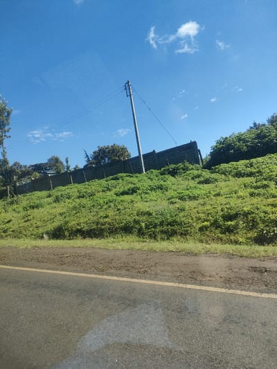 The image shows a scene from a road in an unidentified location within a municipality ward in Kenya. The viewpoint is from a road, with a yellow line marking the edge. To the right is a grass-covered embankment. A grey block wall runs along the top of the embankment, with greenery visible behind it. A single telephone pole stands near the wall. Above, the sky is blue with a few small clouds.