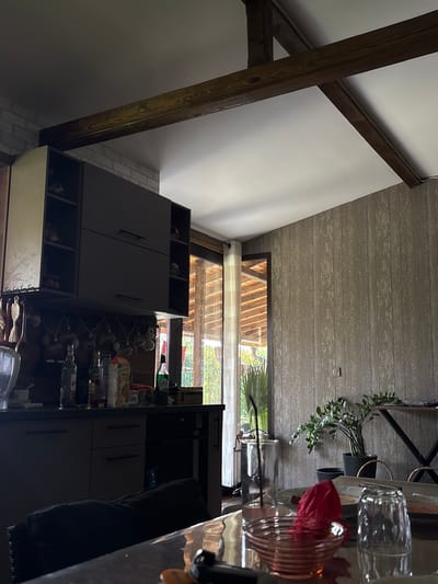 The image depicts the interior of a kitchen and dining area with rustic architectural elements. Visible are dark wood beams against a white ceiling, and a brick wall on the left side of the frame. A set of dark grey kitchen cabinets are mounted on the wall. Below the cabinets, a countertop holds various bottles, including what appears to be a carton of orange juice and a green bottle. A glass and a pink cocktail are on a table in the foreground, alongside a red crumpled napkin and another glass. A green plant in a black pot is situated on the right side of the image. The background reveals a doorway with a sheer curtain, leading to an outdoor area with visible wooden structures. The scene is set in Viden, Bulgaria, suggesting a residential or possibly a small establishment setting. There are no individuals present in the frame.