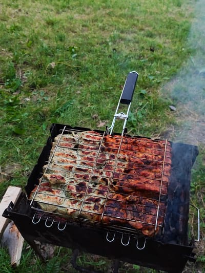 In the city of Sosny, Ukraine, a portable metal grill with a black handle is positioned on the ground. The grill contains a wire rack filled with two types of meat. The left side of the rack holds strips of meat marinated with herbs and spices, appearing lighter in color. The right side features strips of meat with a reddish-brown, well-cooked appearance. Smoke emanates from the right side of the grill, indicating active cooking. The grill is situated on a patch of green grass with some dry leaves and twigs scattered around. In the background, more green grass and a hint of dry vegetation are visible. The scene suggests an outdoor cooking activity.