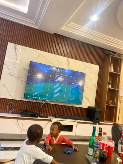 This image captures a modern interior, likely a living room or entertainment area in a home, potentially in Adenta Municipality, Ghana. The main focal point is a large flat-screen television mounted on a wall featuring a striking white marble-patterned slab. The wall behind the marble is covered in horizontal wooden slatted paneling. Above the television, a long, dark wooden shelf is visible, holding decorative items such as vases and gold-colored ornaments.
On the television screen, a video game is being played, displaying a football match scoreboard: "REAL MADRID 0 - 3 MAN CITY." Below the score, game menu options are visible, including "TACTICS & CUSTOMIZATION," with further sub-menus like "Formation," "Roles," "Instructions," "Attacking," "Midfield," "Defending," "General," and "All Tactics." A game controller or console (what appears to be an Xbox Series X and an antenna) and a sound system (speaker and soundbar) are placed near the television.
In the foreground, to the right, two young children are partially visible. One child, wearing a red long-sleeved top, is leaning on a dark wooden table, holding a mobile phone and looking towards the table or television. The other child, wearing a white top, is only visible from the top of the head, also at the table. On the table, several bottles are present, including "Coca-Cola" and "PERRIER," along with other plastic and a distinctive green glass bottle, possibly sparkling wine or champagne.
The ceiling features contemporary recessed spotlights and a linear light fixture, contributing to the bright interior lighting. The overall scene suggests a casual gathering or family time, with children present while a game is being played.
