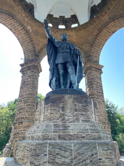 The image shows a low-angle shot of the Kaiser Wilhelm Monument in Porta Westfalica, Germany. The monument features a large bronze statue of Kaiser Wilhelm I, dressed in military attire and holding a sword, standing on a stone pedestal. The statue is situated within a stone arcade structure with arched openings, providing a view of the sky and surrounding trees. The monument is constructed from large stone blocks, and the statue itself is dark, weathered bronze. The sky is clear and blue, indicating a sunny day. Lush green trees are visible in the background, suggesting a natural setting.