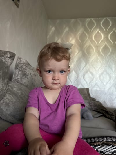 A toddler with blonde curly hair and blue eyes is sitting on a sofa in a room. She is wearing a purple t-shirt and pink leggings with a flower pattern. The sofa is gray with patterned cushions. Behind the child, a wall is visible with a textured wallpaper featuring a repeating organic pattern. The room appears to be indoors, possibly a living room, with soft lighting. The specific location in the city of Beryozovka, Russia is not discernible from the image itself.