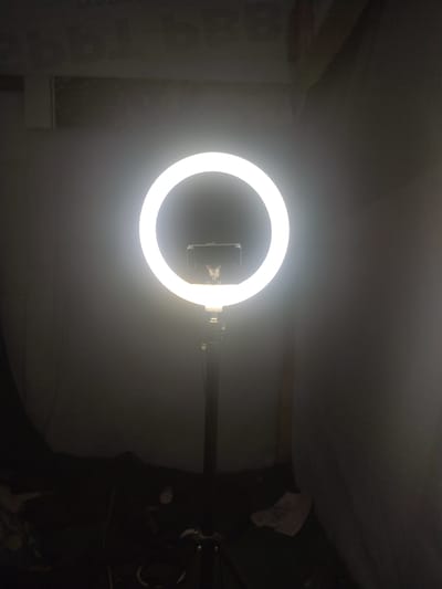 A ring light, illuminating a smartphone holder, is mounted on a tripod. The backdrop consists of dark, draped fabric and a glimpse of a light-colored surface. The bottom portion of the image reveals a cluttered floor with various items, including what appears to be a black bag or garment. The setting is dim, with the bright ring light serving as the primary light source. No people are visible in the scene. The location is not specified to be in Neuheun, Indonesia.