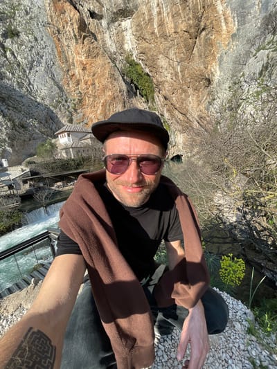 An adult male is captured in a selfie, crouching on a light-colored, rocky ground overlooking the Blagaj Tekke in Dračevice, Bosnia and Herzegovina. The man, with a beard and smiling expression, wears a black baseball cap, purple-tinted aviator sunglasses, and a black t-shirt, with a brown fleece jacket draped over his shoulders. A black geometric tattoo is visible on his left forearm, which extends to hold the camera. In the background, to the left, are the distinct white Ottoman-style buildings of the Blagaj Tekke monastery, situated at the base of a massive limestone cliff. Below these structures, the clear, turquoise Buna River flows, forming small waterfalls. To the right, the river extends towards a dark cave entrance at the cliff's base, with bare tree branches and sparse green foliage visible along the banks. The towering, light-colored cliff face, showing natural reddish-brown and grey formations, dominates the upper background under a clear sky. Strong sunlight illuminates the scene, casting shadows.