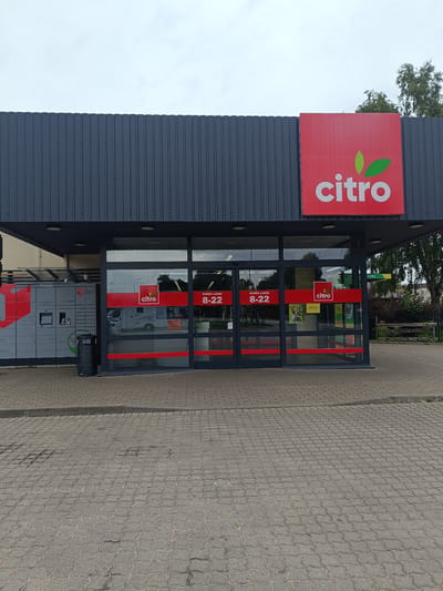 The image shows the exterior of a "Citro" store in Skrunda, Latvia. The store has a modern, dark gray facade with a large red sign displaying the "Citro" logo and name. The entrance features large glass doors with red accents and clear signage. Inside the store, one can see a white RV and various promotional materials. To the left of the entrance is a DPD parcel locker system. The ground in front of the store is paved with interlocking concrete blocks. The sky is overcast. There are no people visible in the scene.
