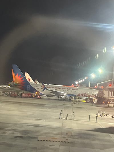 The image depicts two Jet2.com airplanes parked at an airport tarmac at night. The airplane in the foreground has "Jet2holidays" written on its fuselage. The tail of this airplane is predominantly white with an orange and blue design featuring a stylized sun. Another airplane is partially visible behind it, with a similar tail design. Several ground vehicles, including what appear to be baggage tugs and possibly fuel trucks, are positioned around the aircraft. Airport lighting poles are visible in the background, illuminating the scene. The sky is dark, suggesting it is nighttime. The scene is set at Alicante–Elche Miguel Hernández Airport (ALC), located in Elx/Elche, Spain.