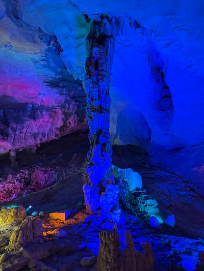 This image shows a section of the Shui Cave scenic area, located in the city of Zhuoshui, China. The interior of a cave is visible, exhibiting various rock formations illuminated with blue, purple, and yellow artificial lighting. The rocks and cave walls display textures and shapes indicative of natural erosion and mineral deposits. Two small figures appear at the bottom of the cave in the lower right portion of the image.