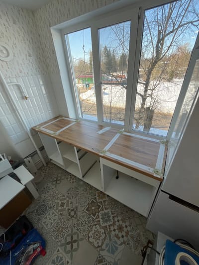 This image depicts an interior room, located in Нарынка, Russia, observed during daytime under clear skies. The room is undergoing renovation or furnishing. The floor is covered with a mosaic of patterned ceramic tiles in shades of brown, grey, blue, and white. The left wall features a lower section of white rectangular tiles, above which is wallpaper with a light background and a subtle leafy pattern. A white radiator pipe with a brass-colored valve extends from the tiled wall.
A large, three-pane window with white frames occupies the central portion of the image, providing a view of an outdoor environment. Outside, patches of snow are visible on the ground, indicating late winter or early spring. Bare deciduous trees and distant evergreen trees are present. A building with a green roof is partially visible in the mid-ground.
Beneath the window, a newly installed wooden-look countertop is visible, spanning the width of the window. Several strips of white masking tape are applied to the countertop surface, and small deposits of a light green substance, consistent with adhesive or sealant, are present at the tape's corners. Below the countertop are white, open base cabinets, some displaying internal shelves. To the far right, a tall, white appliance, possibly a refrigerator, is partially visible. On the floor in the lower-left foreground, a blue bag containing items and additional white cabinet components are present. No individuals are visible within the scene.