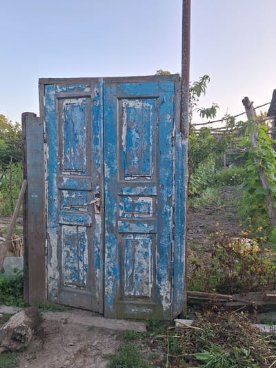 The image shows a weathered, blue wooden double door set within a rustic outdoor environment. The paint on the doors is peeling, revealing older layers of paint and the bare wood underneath. The doors have a simple design with rectangular panels. A metal door handle is visible on the right-hand door.
To the left of the doors, a rough wooden post stands upright, and a long, wooden tool, possibly a rake or shovel, leans against it. Behind the doors, a garden or yard is visible, with plants, vines, and trellises suggesting cultivation. A metal pole stands near the doors, and a wooden fence or structure is present further back. The ground in front of the doors is a mixture of dirt and some green vegetation. The overall scene evokes a sense of neglect or disuse, but with the underlying signs of a functional outdoor space.
The image is located in the city of Nabran, Azerbaijan, a resort town known for its natural beauty and coastal landscapes. The setting depicted is consistent with the rural or semi-rural character that can be found in areas surrounding such towns. The presence of a garden and the rustic nature of the door suggest a private property, possibly a residence or a shed within a larger estate. The time of day appears to be either early morning or late afternoon, given the soft lighting and the clear sky.