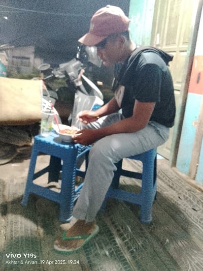 The image shows a young man wearing a black t-shirt and a brown baseball cap. He is sitting on a blue plastic stool and eating from a white bowl with a red plastic spoon. There is a glass of liquid and a red straw on the stool next to him. He is sitting in front of a partially obscured building. The floor is brown and the surrounding environment is poorly lit, suggesting nighttime. The location is near the coordinates -6.422, 107.996 which is in Kecamatan Kroya, Indonesia.