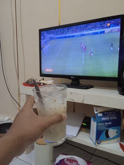 This image captures an indoor scene, likely a residence in Brangsong, Indonesia, where a person is engaged in leisure activities. The foreground shows a light-skinned hand holding a clear glass containing a light, opaque beverage with ice and a metal spoon. The glass is adorned with patterns resembling stars and Islamic lanterns. In the midground, a black LG television displays a soccer match in progress, featuring players in red and white jerseys on a green field with a white goal. Overlays on the screen include "INDOMILK," "LANGSUNG," and "REPL." The television rests on a white, weathered wooden shelf. To the left of the TV on the shelf, a red tray holds various metal clips and small items. Below the television, on a lower shelf, a stack of white papers, a small black device with green digital numbers (likely a clock or timer), and an open blue and white box labeled "FACE MASK" (indicating 50 disposable 3-ply masks) are visible. A black speaker is partially seen to the right of the TV. The background consists of a plain, light-colored wall with vertical panel lines and various black electrical wires running along it. A yellow cable extends vertically in the upper right. On the floor or a lower surface, a white ceramic container with a wooden lid and part of a pink and white plastic bag are visible.
