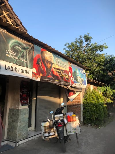 The image shows an outdoor scene in the city of Putat, Indonesia. The foreground features a motorcycle parked on a paved surface, loaded with various containers, including what appear to be condiment bottles. A person's legs are visible on the left side of the image, partially obscured. Behind the motorcycle is the facade of a building with a corrugated metal roof supported by wooden beams. Several large banners advertising cigarette brands are displayed along the front of the building. The banners are colorful and feature images of people and text in Indonesian. Further back, a wall made of red brick is visible, with a green, leafy bush growing in front of it. The sky above is clear and blue. The overall impression is of a street scene in a tropical urban environment, likely during daylight hours.