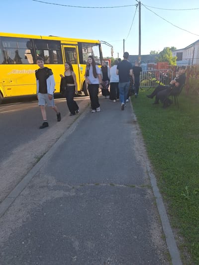On a paved path next to a road, a group of people are walking. On the left, a bright yellow bus is parked. A young man in a black and white jacket and grey shorts is walking towards the camera. To his right, a young woman in a black tank top and dark pants is walking. Another young woman in a white t-shirt and black pants is walking behind them. Further back, several other individuals are walking away from the camera, including a man in a dark t-shirt and jeans. To the right of the path, a grassy area is visible, with a fence and a building in the background. Two people are sitting on chairs in the grassy area, one of whom appears to be wearing a dark uniform and a cap. The sky is clear and blue. The scene appears to be in a residential area.