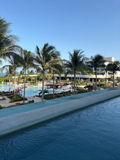 The image captures a view of a resort in Punta Sam, Mexico, from an elevated vantage point, likely an infinity pool. In the foreground, a section of the turquoise water of the infinity pool is visible, with subtle ripples on its surface. A textured, light gray concrete barrier separates the pool from the resort grounds.

Beyond the barrier, a large, multi-level resort building with white and beige facades and balconies dominates the background. Numerous palm trees are scattered throughout the scene, with their fronds casting shadows. The resort grounds feature multiple swimming pools, some with loungers and umbrellas, and a sandy area that appears to lead to the ocean. Several thatched-roof structures are interspersed among the greenery.

People are visible in and around the pools, some swimming and others relaxing on loungers. A pathway or deck area connects different sections of the resort, and tables and chairs are set up in an outdoor dining or lounge area. The sky is a clear, bright blue, indicating a sunny day. In the far distance, the ocean is visible, merging with the horizon. The overall scene depicts a tropical resort setting.